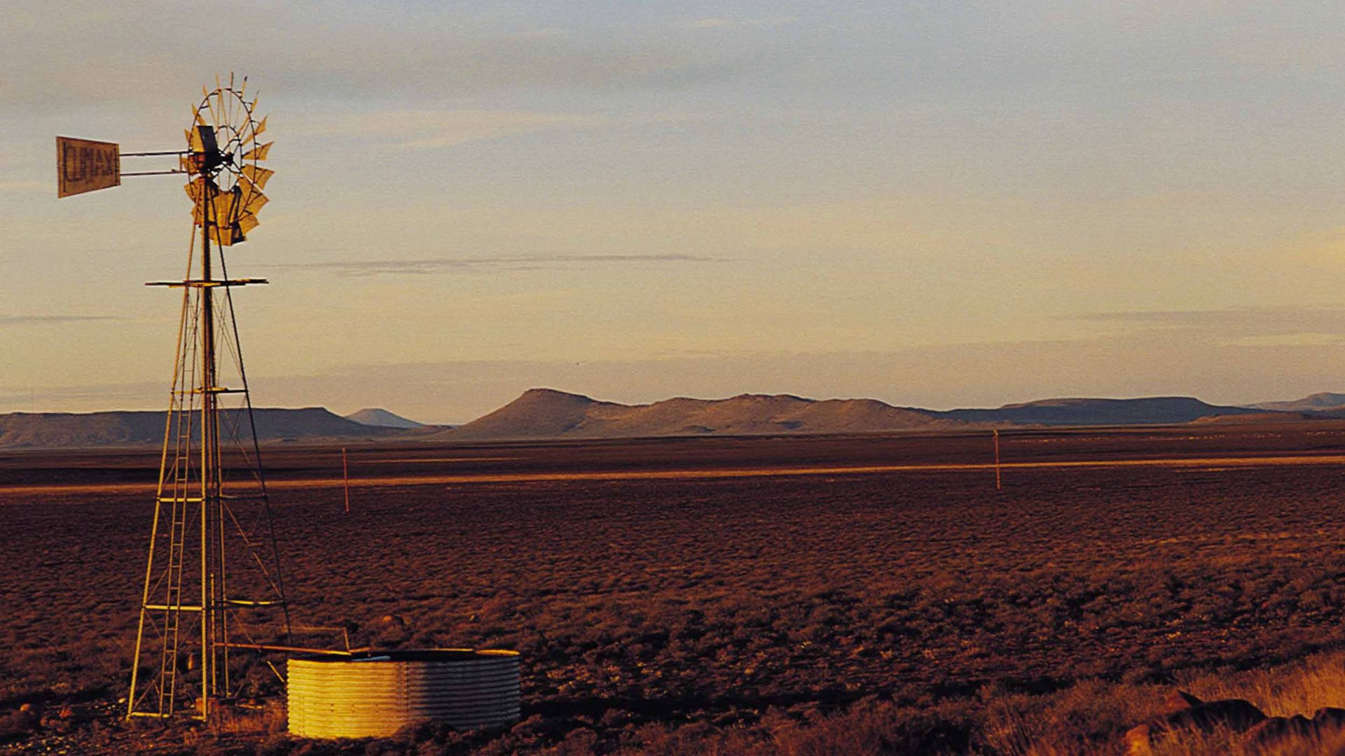 A Karoo windpump and water tank at golden hour, flat-topped koppies on the horizon